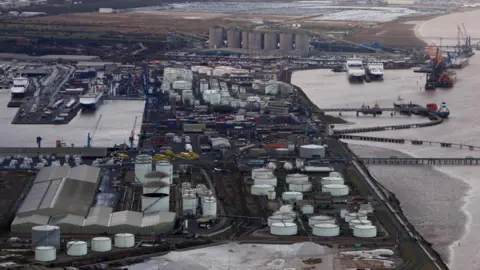 An aerial view of Immingham docks. Four ferries are parked, two either side of a series of energy plants. Cooling towers are in the distance, along with a number of cranes