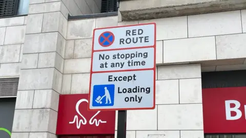 A white road sign with red borders on a street in Brighton, it reads "red route, no stopping at any time, except loading only"