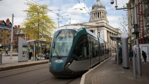 Getty Images A modern light grey and green tram is seen at a stop in the centre of Nottingham. Behind the tram is the city's council house