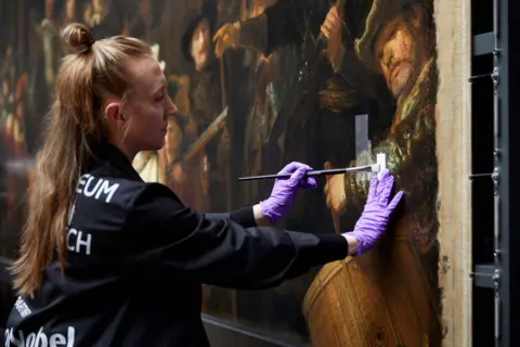 Courtesy of the Rijksmuseum A woman with long blonde hair in a ponytail is wearing black protective clothing and purple latex gloves as she carefully removes the varnish from a section of The Night Watch