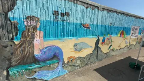 John Nurden A seafront mural featuring a mermaid, seal, crab, boat and children playing in sand.
