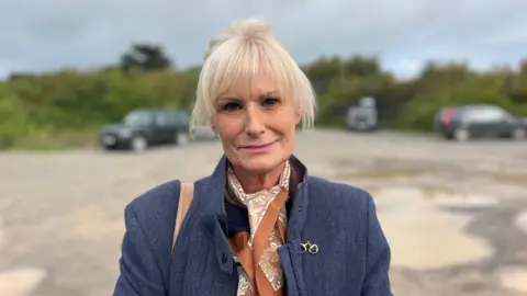 Catherine Jones who is wearing a tweed smart jacket, brown and white cravat, has blonde hair and looking at the camera