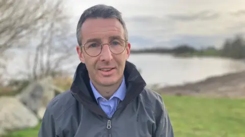 Andrew Muir wearing smiling wearing a grey coat and blue shirt and glasses. His hair is short and grey. 