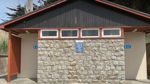 Front view of a public toilet building with a stone central wall, cream side walls, and a dark wooden pitched roof. Small rectangular windows run beneath the roofline. Blue signs on either side indicate “Gents Hommes” and “Ladies Dames,” with a central sign reading “Côtíl du Grouin Toilets.”