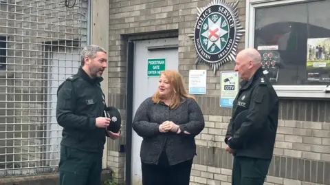 Naomi Long stands in between two senior police officers outside of Lurgan police station. They are all in conversation. The station is a grey brick building, with a metal caged wall on the left hand side.