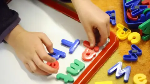 A pair of a child's hands are putting magnetic primary-coloured plastic letters on a small board.