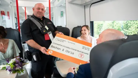 Transport for Wales A ticket officer for Transport for Wales holds the enlarged train ticket offering congratulations. Bethan sits at a tabled seat in a train carriage.