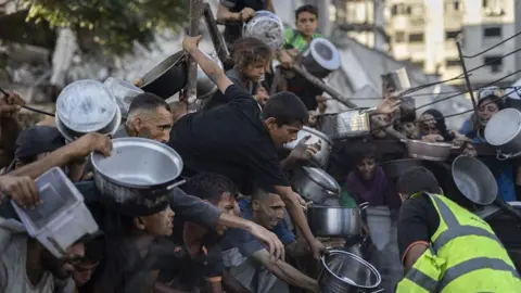Anadolu via Getty Images Palestinians hold out pans at a charity kitchen in the al-Rimal neighbourhood of Gaza City (11 July 2025)