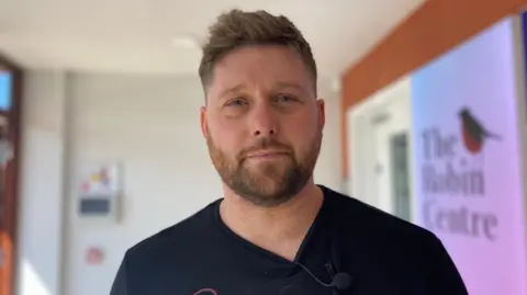 A man has short brown hair and a beard. He is wearing a black top and is standing in front of an orange and white wall. There is a blurred image of a robin and the words The Robin Centre on one wall.