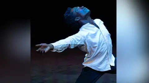 Nahum McLean/Eloise Frey Male dancer, Nahum, wears a white shirt and dark trousers on stage underneath a blue stage light. His arms are wide open and he is lunging as he turns his head to the ceiling.
