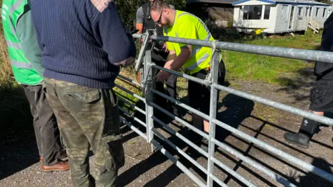 A man in a high visibility jacket and a man in a black stab-proof vest bolt a steel five-bar gate while two men look on.