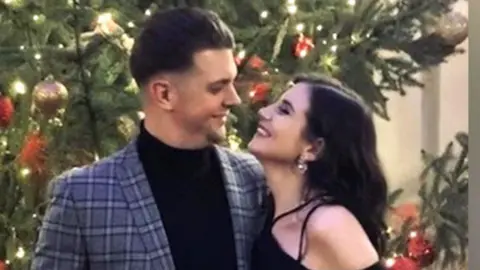 Family photo Jack Hurn and girlfriend Alex Jones looking at one another and smiling, they are standing in front of a Christmas tree