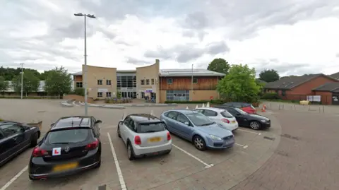 Google Line of cars parked in a car park with a community centre that is made from pale stone and wood behind them 
