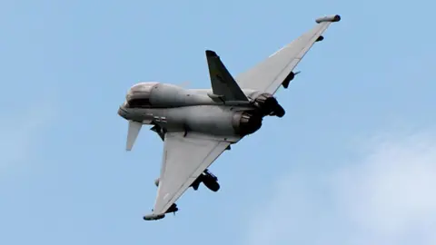 A grey fighter aircraft is seen in the distance flying against a blue sky 