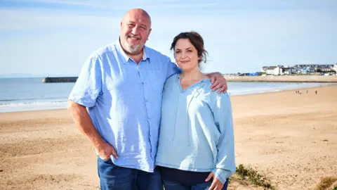 ITV Cymru Wales / BBC Cymru Wales Jones and Speirs standing together with Porthcawl beach behind them. It is a sunny day and they are both smiling looking at the camera. Speirs, on the left, wears a blue linen shirt and navy jeans. He has his arm around Jones, who is wearing a blue blouse and navy jeans.