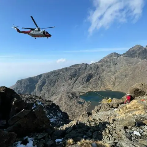 David Sked A red and white coastguard helicopter hovers above where rescue team members help to arrange for the casualty to be winched onboard. 