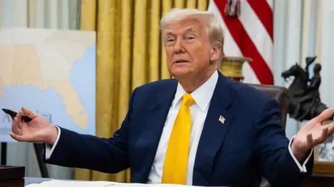 Donald Trump, sitting in the Oval Office, spreads his arms and hands besides him. He is holding a pen in his right hand