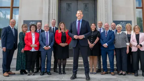 Welsh government The first minister and cabinet ministers stand on steps in front of a grey building with a brown door.
