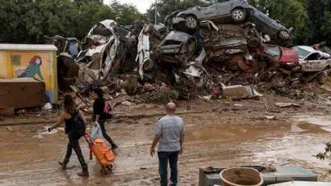 Getty Images A man stands and looks at a pile of flood-damaged cars. car is being moved from the road