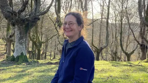 Helen Read Helen Read sits in a forest of spaced out ancient trees, with dappled sunlight on the mossy ground.