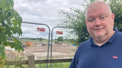 A man with a grey crew-cut hairstyle and a blue polo shirt is standing in front of his garden fence. Behind, there is a metal fence and the foundations of a number of homes.