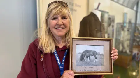 A woman with blonde hair and a maroon top holds up a framed picture of a horse and foal