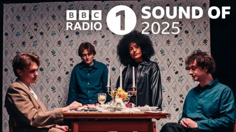 Denmarc Creary Promotional photo of the indie band English Teacher, taken in a mock-up of a suburban living room. The logo for BBC Radio 1's Sound of 2025 is superimposed onto the background.