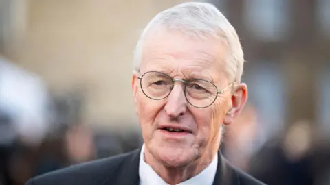 PA Media A close-up of Hilary Benn. He is an older man with short white hair. He is wearing a pair of circular metal glasses. He is wearing a dark suit jacket and a white shirt.
