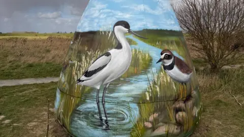 Nathan Murdoch A close up of a buoy in a nature reserve featuring paintings of two birds near a stream. 