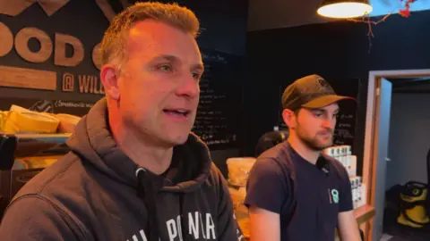 A man with short hair and a dark hoodie sits next to a man with a blue T-shirt and a dark cap