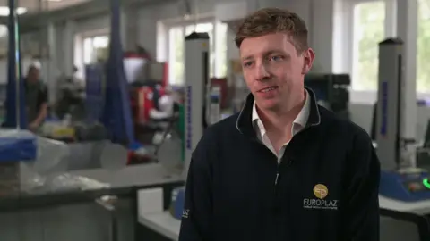 Rory is wearing a black quarter-zip jumper with the Europlaz logo on it. He is sitting in a laboratory-type room with machines visible in the background. He has short, fair hair. 
