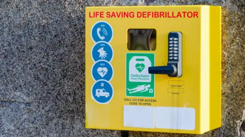 Getty Images Yellow defibrillator box attached to pebble-dashed wall. It reads "life saving defibrillator" and has a code lock.