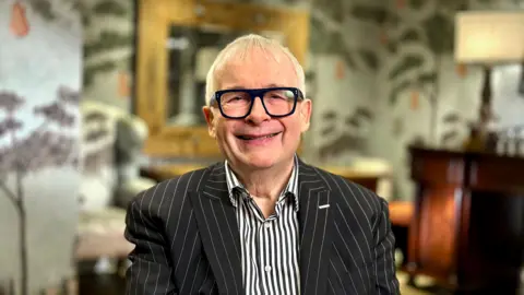A man with short grey hair wears large black glasses with clear, colourless lenses. He wears a black and white pinstripe suit with a black and white striped shirt. Behind him is an out-of-focus grey wall with purple tree's on it.