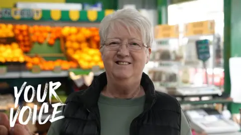 An older woman smiles while standing in a greengrocers.