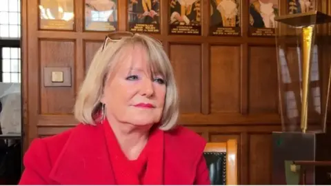 A woman with blonde hair and sunglasses on her head sits in frong of some wood pannelling with what looks like pictures of previous mayors behind her. She is wearing a red jacket.