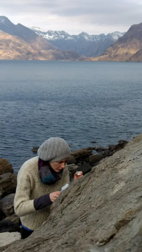 Dr Elsa Panciroli Dr Elsa Panciroli is busy inspecting a rock formation on an area of coastline with mountains on the opposite shore. She is wearing a thick woollen jumper, a scarf and woollen hat.