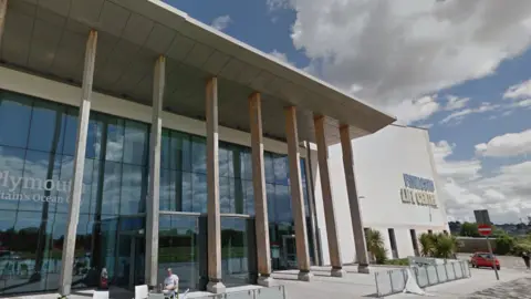 The outside of Plymouth Life Centre. It is a large glass fronted building with seven pillars propping up an extended solid awning. There is cloudy sky in the background of the image.