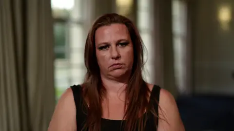 A woman wearing a black tank top and long reddish brown hair sits and stares at the camera.