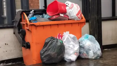 A large orange wheelie bin with bags of rubbish overflowing and three bin bags placed outside it filled with rubbish.