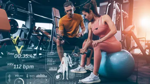 Getty Images A woman in the gym with a personal trainer showing a her a phone. The screen is overlayed with data.
