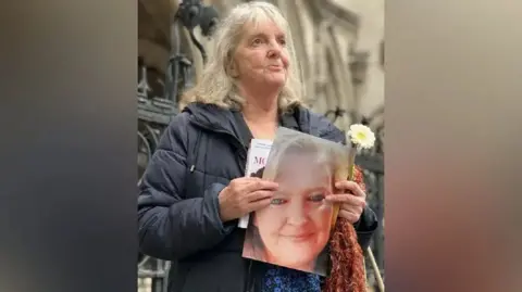 Joy Dove stands outside the Court of Appeal in London. She has shoulder-length grey hair and is wearing a blue coat and is holding a photograph of her daughter Jodey Whiting and a yellow and white flower