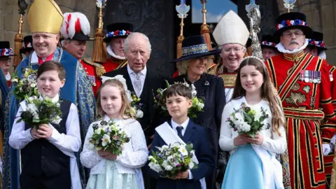 Reuters King Charles and Queen Camilla posing for a group photo outside St. Asaph Cathedral. Four children can be seen stood infront of the royals, each holding flowers.
