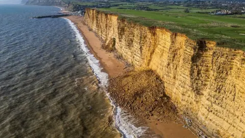 Grass-topped cliffs with golden face and large pile of rocks sprawling across the beach with waves lapping