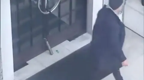 CCTV image showing a cylindrical object propped upright against the glass doors of a building entrance, with a small green lighter lying nearby, as a man in a dark coat walks away.