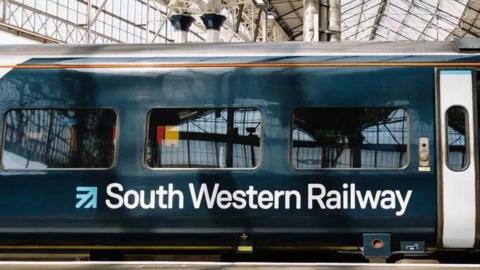 Dark blue with white logo writing South Western Railway train stopped at a station with sliding door open