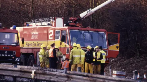 A fire crew are gathered together in front of a fire engine. They are parked on the side of the road with an embankment to the side of them. They are wearing hi vis and are covered in what looks to be oil. 