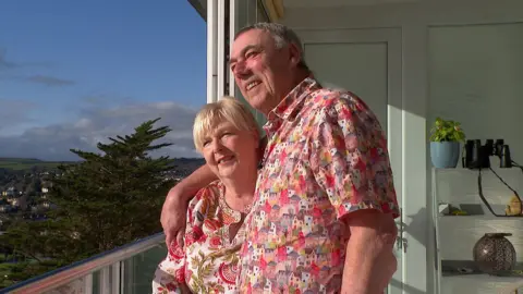 BBC Bob and Lesley Perrin with their arms around each other wearing bright pink tops.  The couple are standing on their balcony smiling looking out at the view.