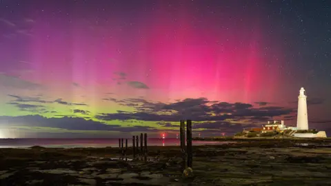 BBC Weather Watchers/Paul Appleby A view across a rocky beach as the tide is out, revealing old tall wooden beams sticking up. In the distance is a white light house which is clearly visible. The sea is far out. The night sky is lit up in green and pink, with lots of stars, as the northern lights are clearly visible.