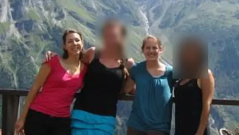 Magali Tassone A group of four people stand together on a viewing platform with a railing, posing in front of a dramatic mountain landscape featuring steep rocky cliffs and a waterfall. Two faces are blurred. One person wears a bright pink top, another a black top with a blue skirt, another a blue top, and another a dark outfit. The scene is outdoors in daylight with rugged green and gray mountains in the background.