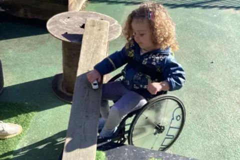Pearson family Grayce Pearson is sitting in a small wheelchair while playing with a toy car on a wooden plank which is set up like a bridge. Another toy vehicle is on the artificial‑grass-covered section of the platform. Grayce has shoulder-length,  curly, blonde hair and is wearing a blue hoodie and grey leggings.